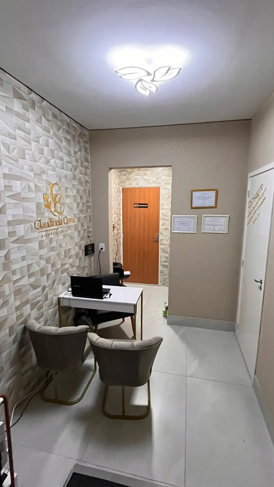 Recepção do consultório da Dra. Claudineia Costa, com o logo dourado na parede de revestimento 3D, mesa e poltronas
